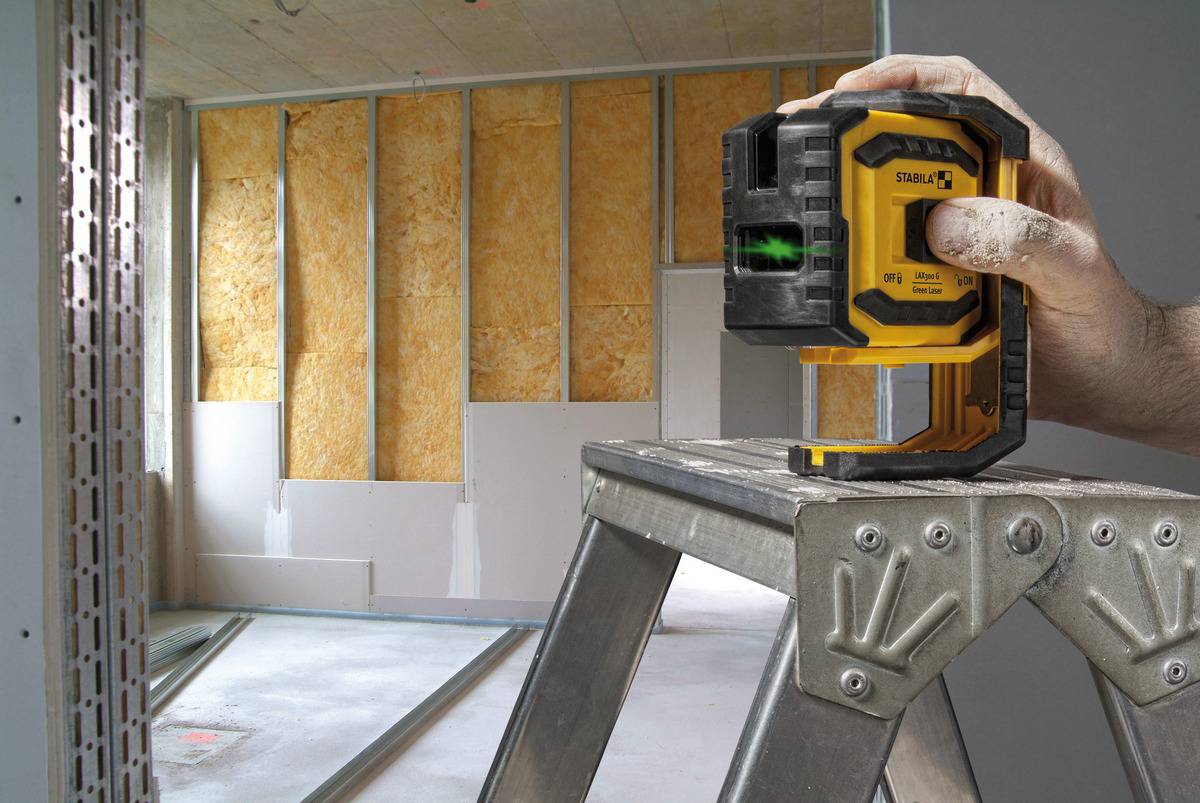 Jedna ręka trzyma laserowy niwelator na drabinie w pomieszczeniu będącym w stanie surowym; w tle widać niedokończone ściany.