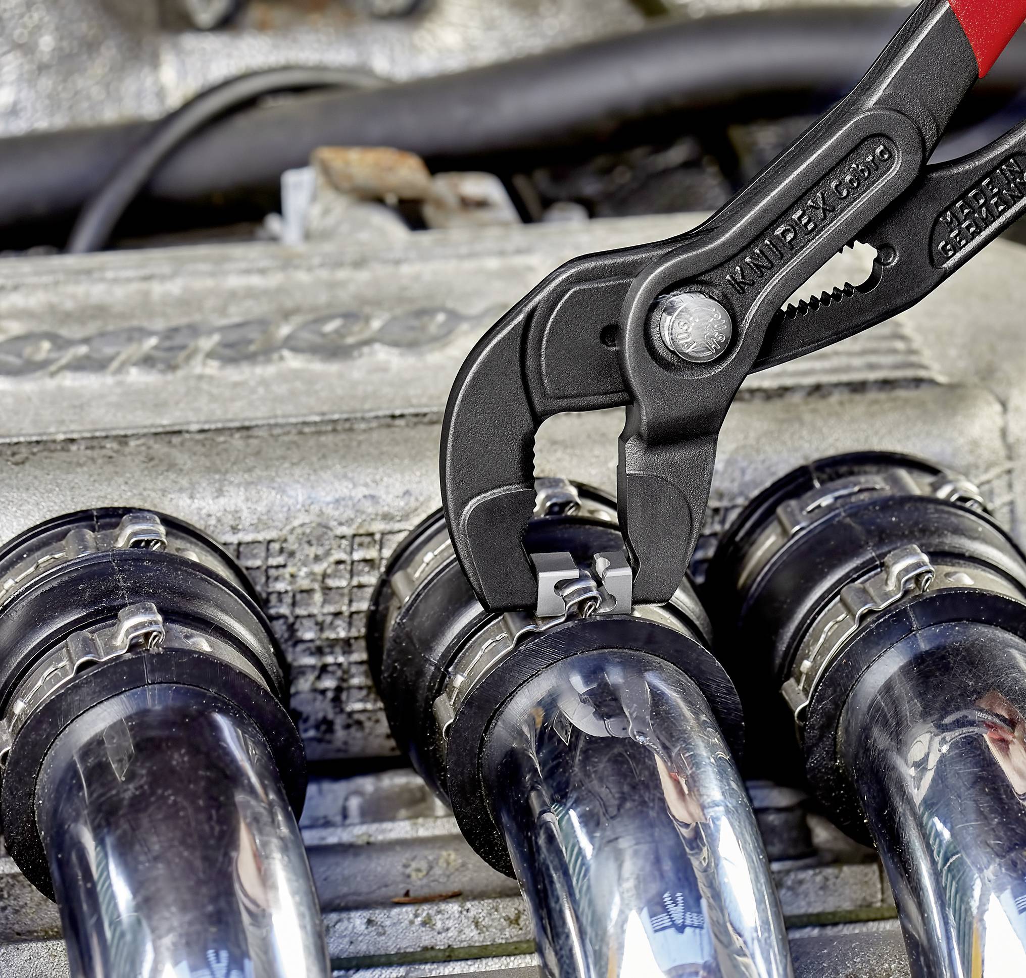 Szczypce chwytają zacisk na metalowej rurze w komorze silnika. W tle widoczne są części mechaniczne.