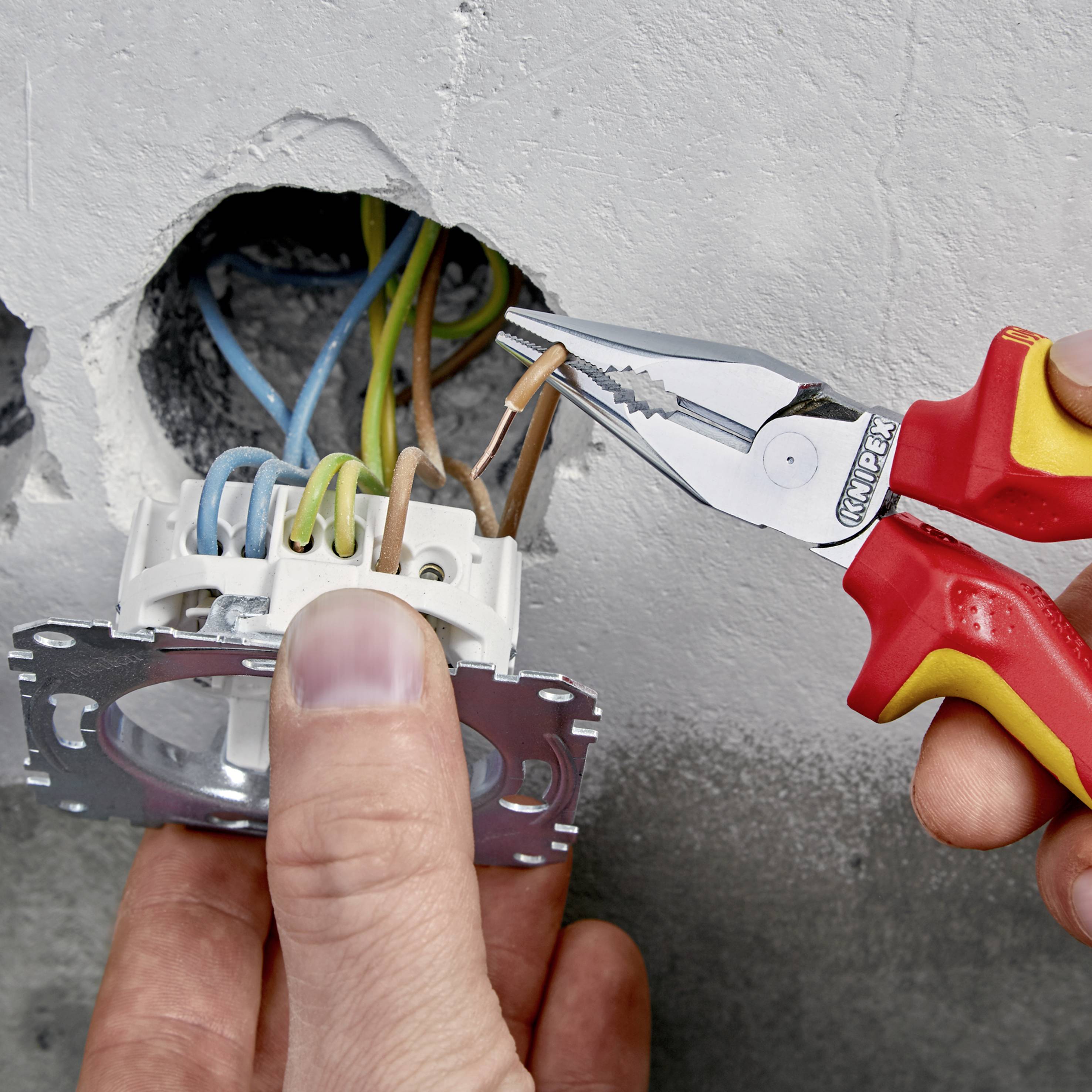 Jedna ręka trzyma szczypce i pracuje przy przewodach elektrycznych w gniazdku ściennym. Druga ręka przytrzymuje gniazdko.