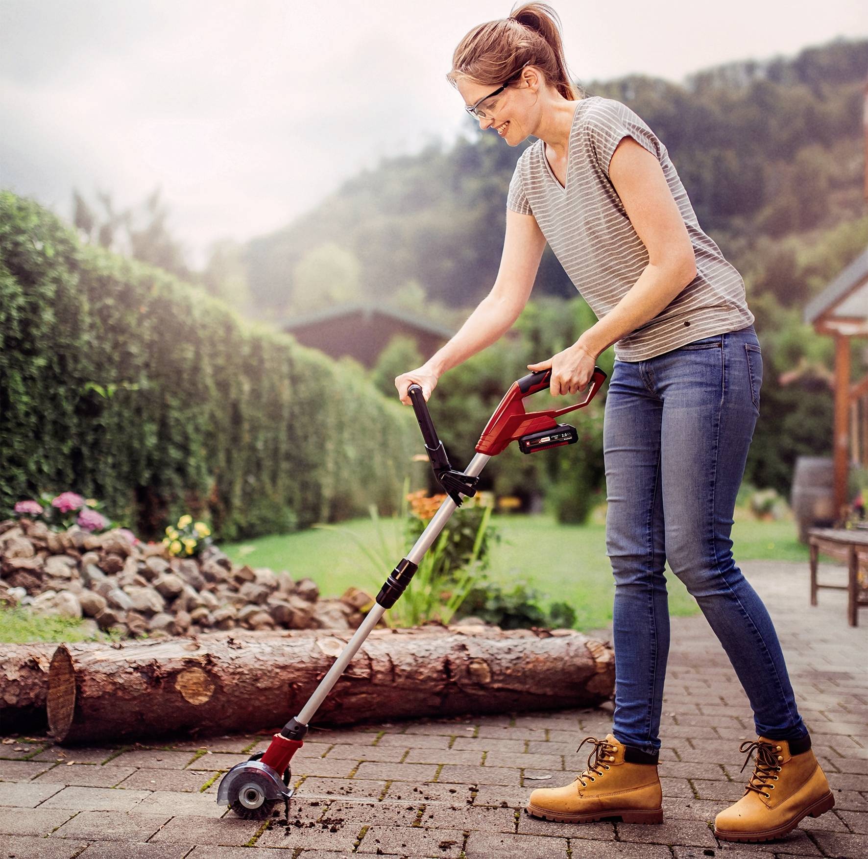 Kobieta używa narzędzia ogrodowego z kółkami do czyszczenia ścieżki między grządkami, otoczona drzewami i łąką.