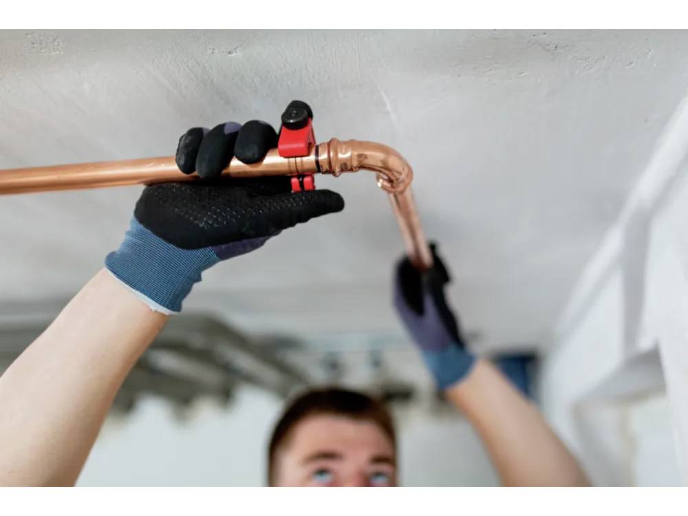 Osoba używająca obcinaka do rur podczas naprawy miedzianej instalacji hydraulicznej pod sufitem, nosząca ochronne rękawice.