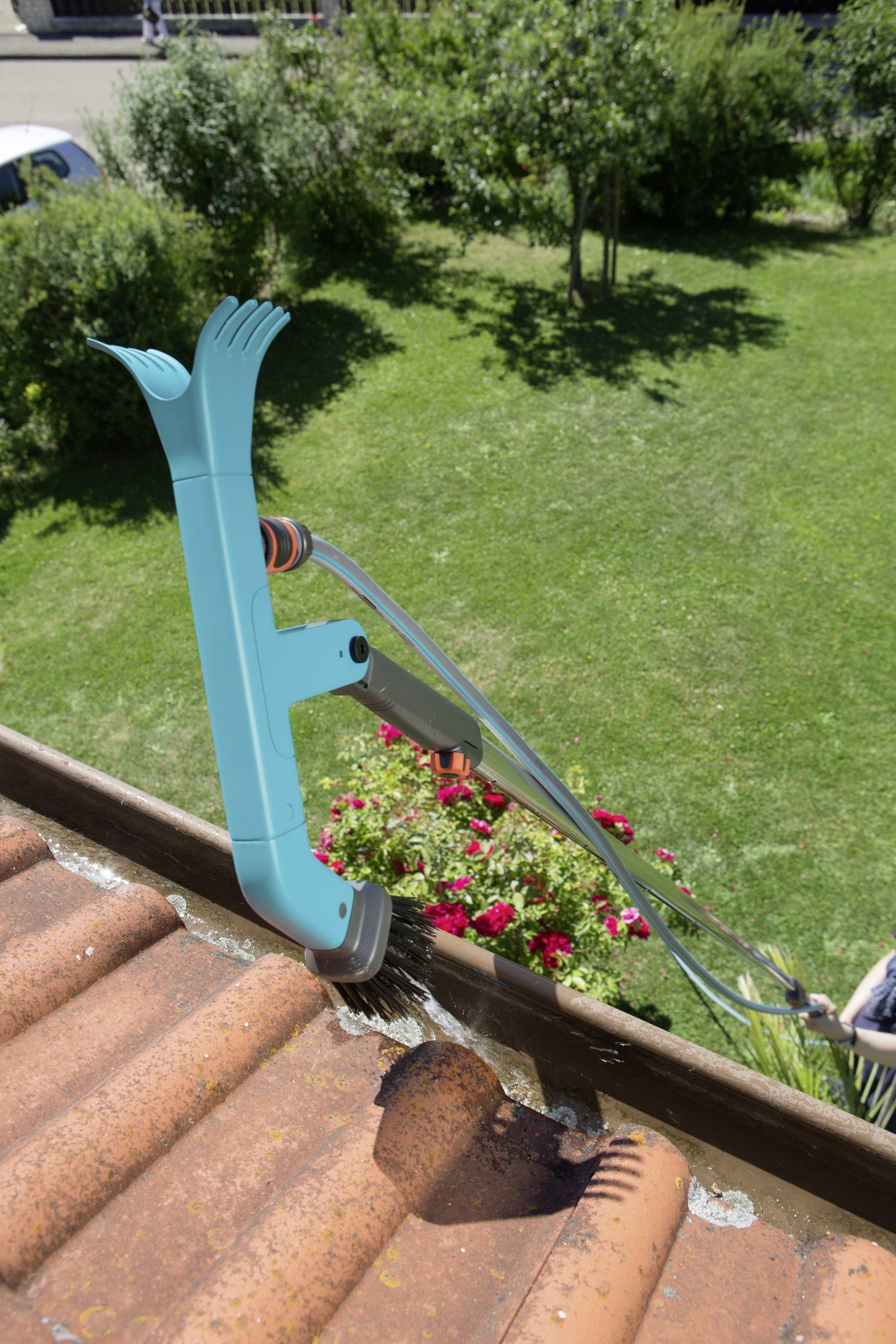 Niebieskie narzędzie czyści porośniętą mchem rynnę na brązowym dachu. W tle widać zielone drzewa i ogród.