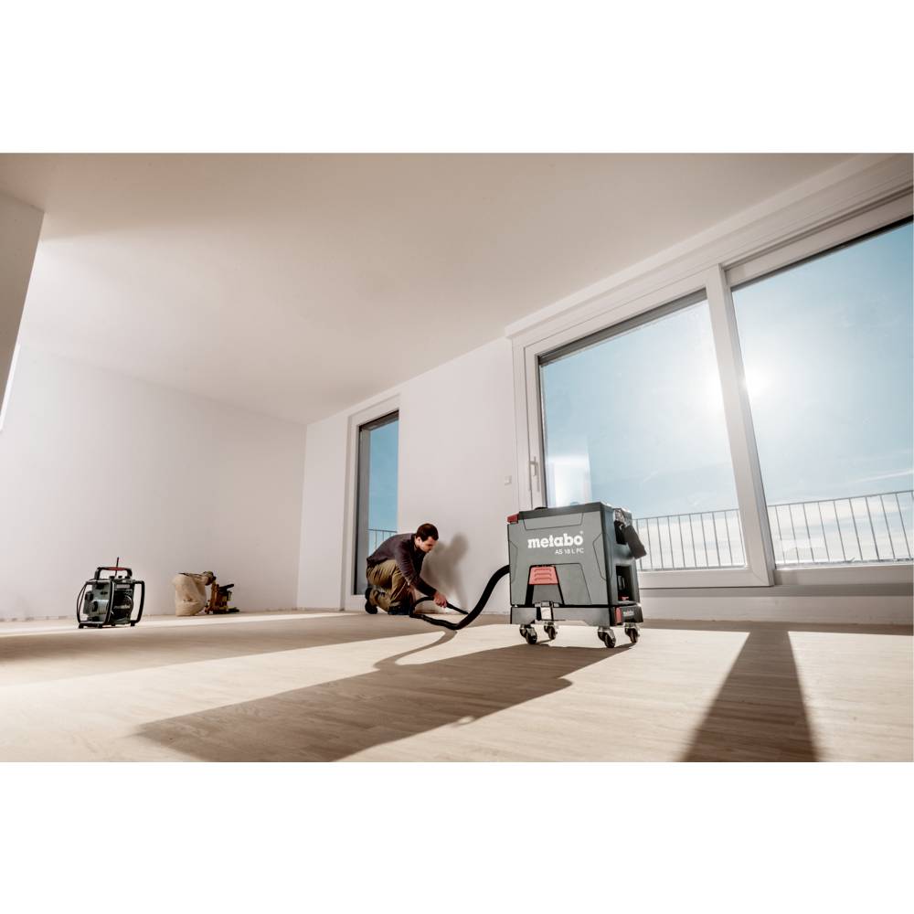Zielony odkurzacz Metabo AS 18 L PC o zwartej, pudełkowatej budowie, wyposażony w kółka, wąż i uchwyt, podkreślający przenośność i przemyślany design.