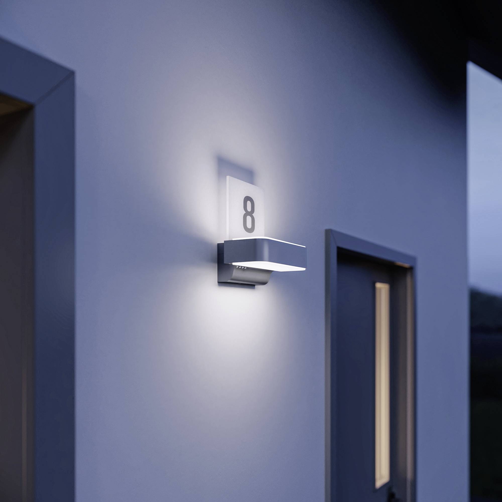 Kinkiet oświetla numer domu 8 na niebieskiej ścianie. Po prawej stronie znajdują się drzwi. Oświetlone otoczenie o zmierzchu.