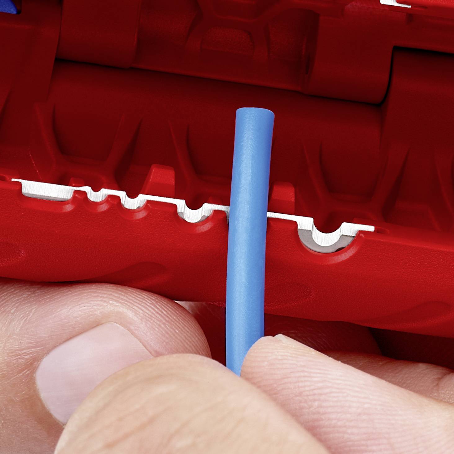 Dwie ręce trzymają niebieski kabel przed czerwono-białym narzędziem używanym do zdejmowania izolacji z przewodów.