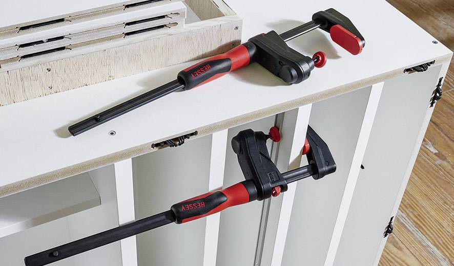 Dwie ścisk stolarskie w kolorze czerwonym i czarnym mocują element mebla w warsztacie.