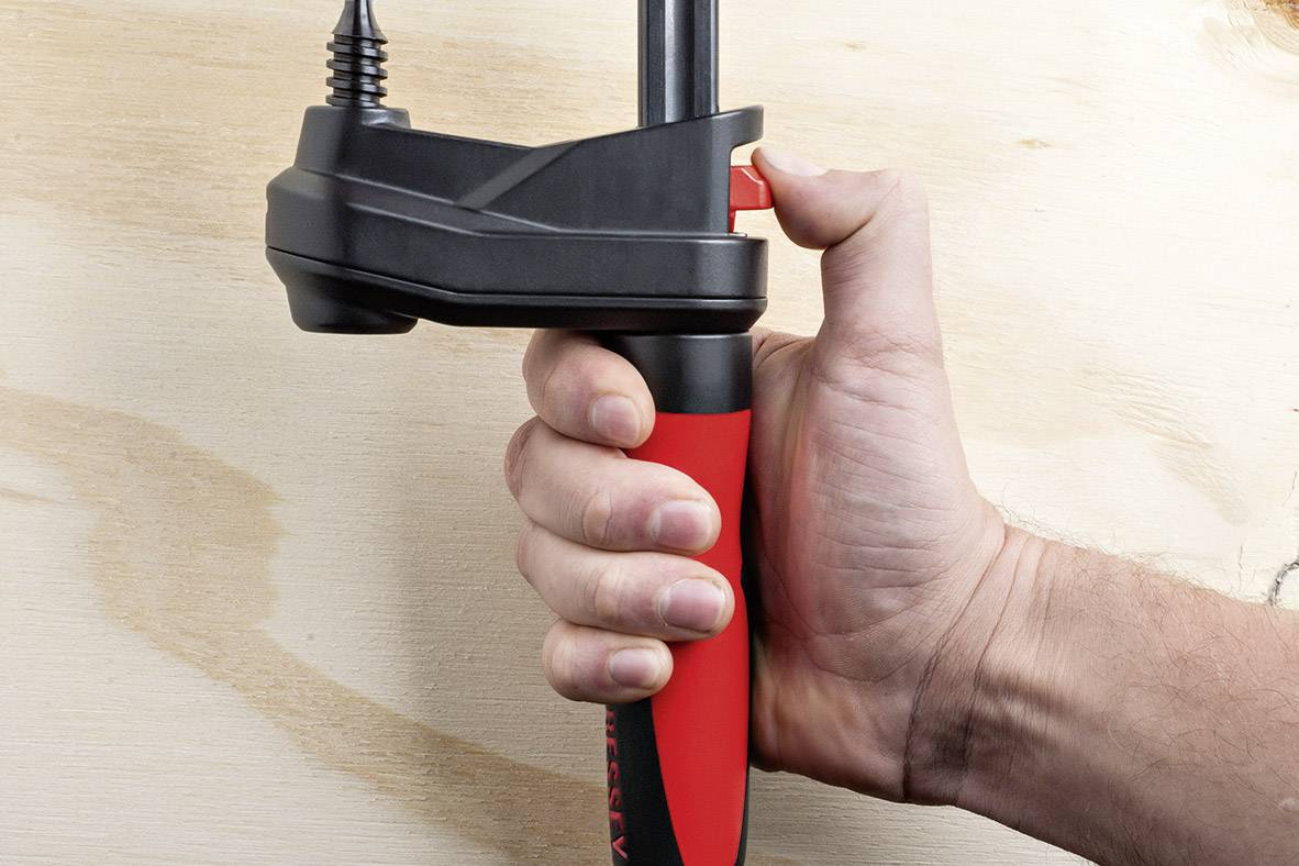 Dłoń trzyma czerwony i czarny uchwyt z czarnym mechanizmem zaciskowym na kawałku drewna.