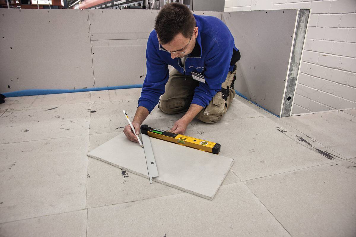 Mężczyzna klęczy na podłodze wyłożonej płytkami i mierzy za pomocą poziomicy i ołówka. Narzędzia leżą obok.