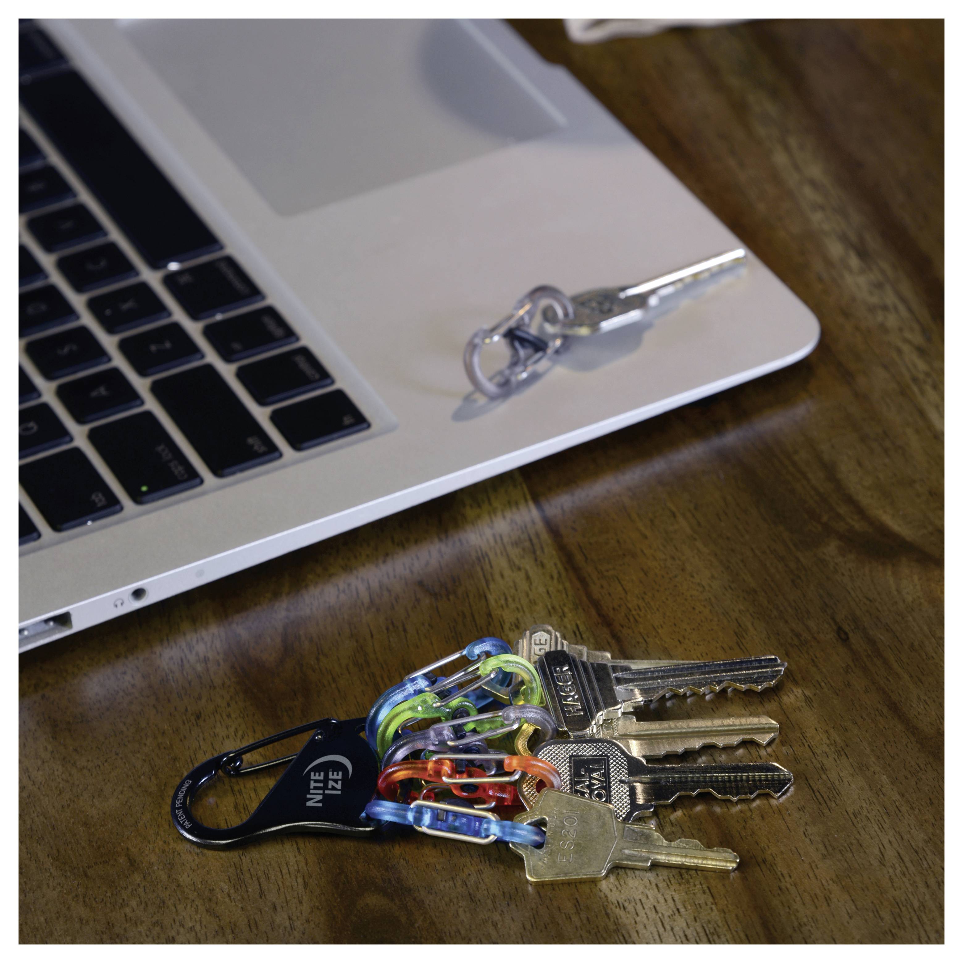 Klucze przypięte do kolorowego kółka leżą na drewnianym stole obok laptopa, a drugi komplet kluczy umieszczony jest na laptopie.