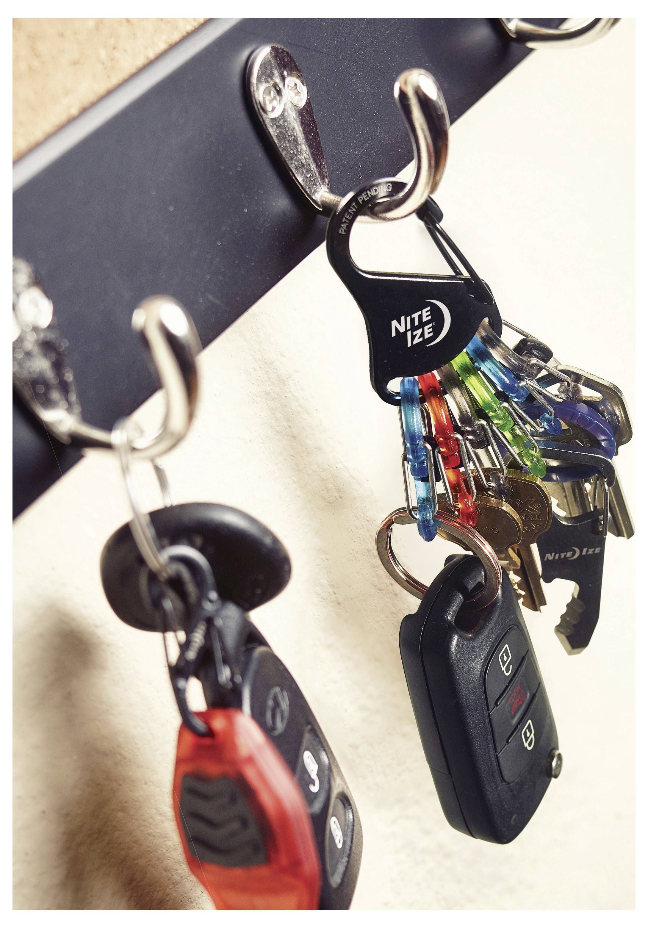 Kilka kompletów kluczy i breloków wisi na metalowych haczykowatych uchwytach na ścianie zamocowanej półce.