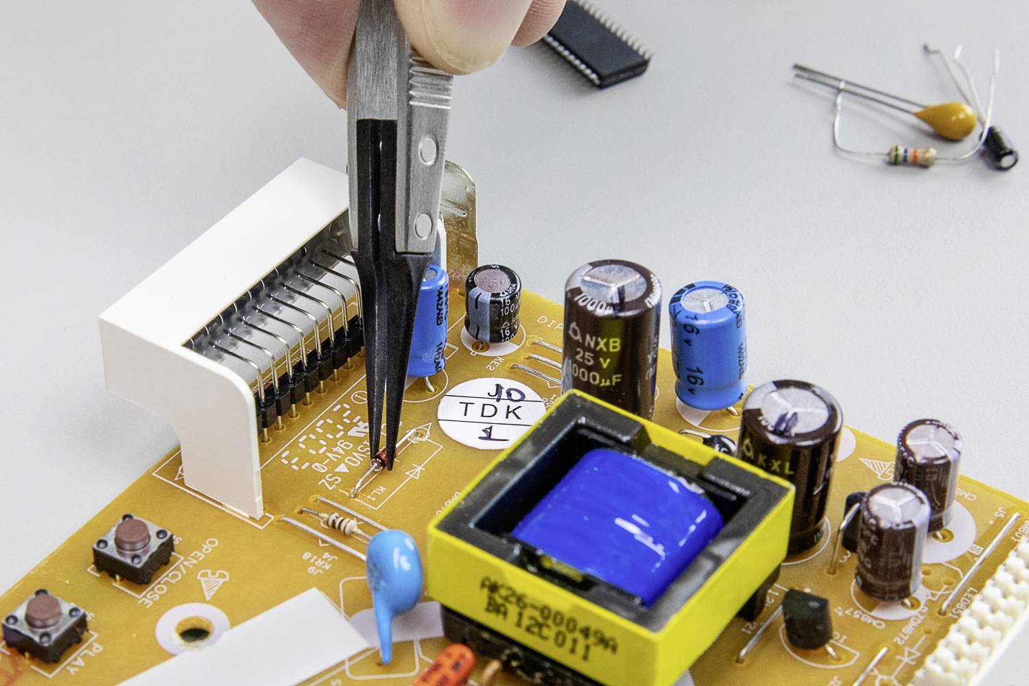 Ktoś trzyma pęsetę, aby naprawić lub zainstalować element elektroniczny na otwartej płytce drukowanej.