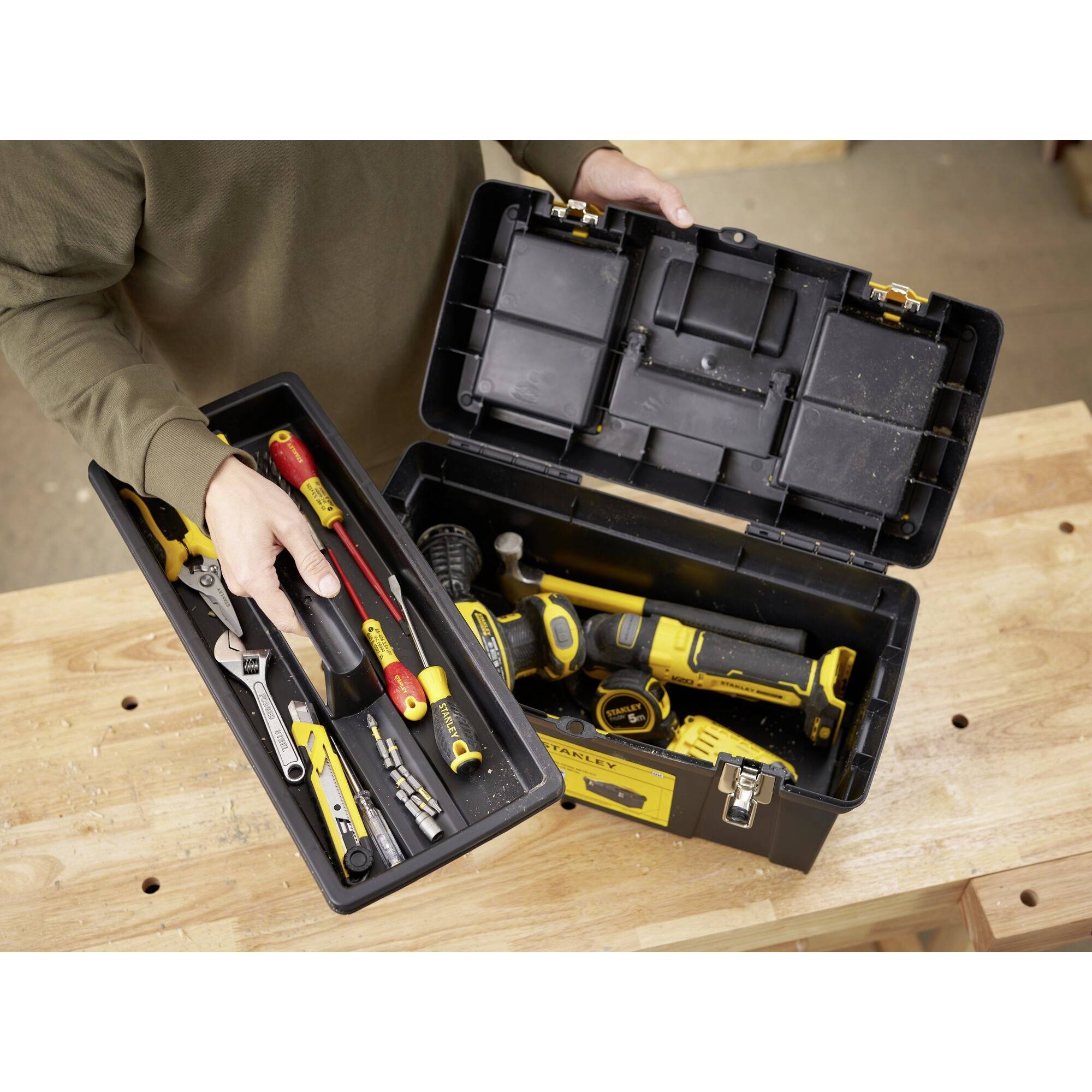 Otwarta skrzynka narzędziowa z różnymi narzędziami ręcznymi, takimi jak śrubokręty, poziomica i klucz nastawny. Jedna osoba trzyma skrzynkę z narzędziami.