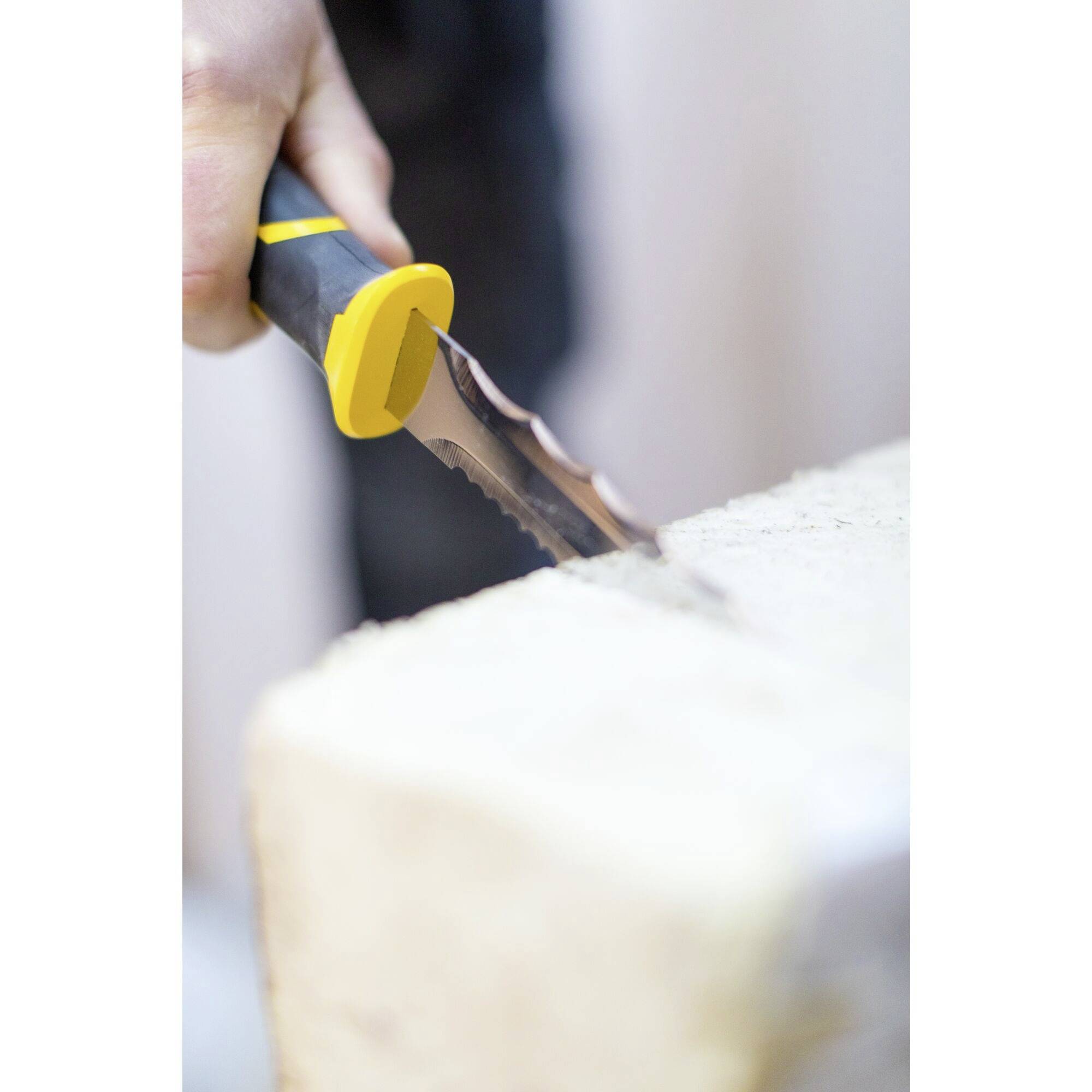 Dłoń używa noża z ząbkowanym ostrzem i żółtą rączką, aby zeskrobać materiał z jasnego bloku.