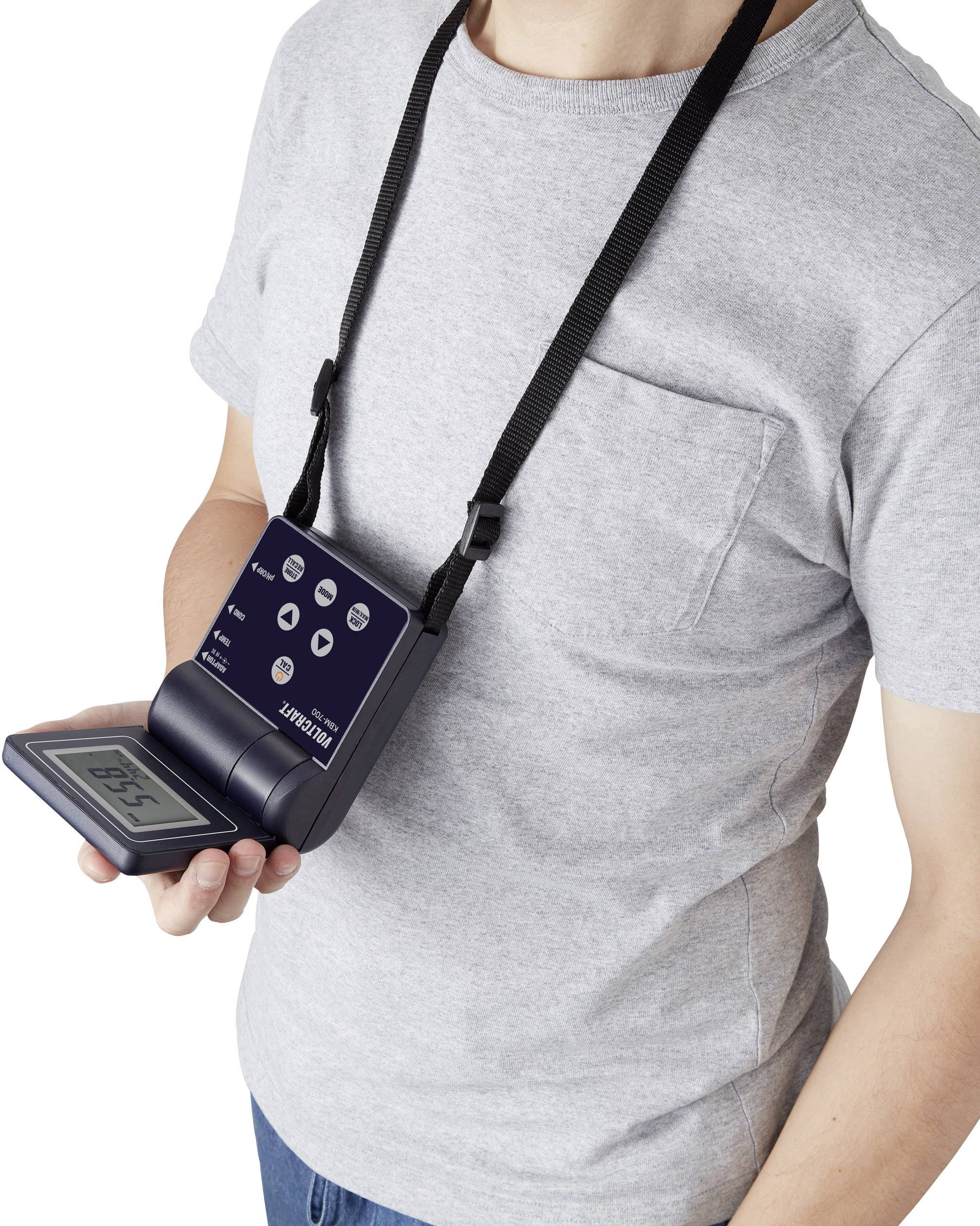 Osoba w szarym t-shircie trzyma przenośne urządzenie elektroniczne z czytelnym wyświetlaczem, zawieszone na szyi.