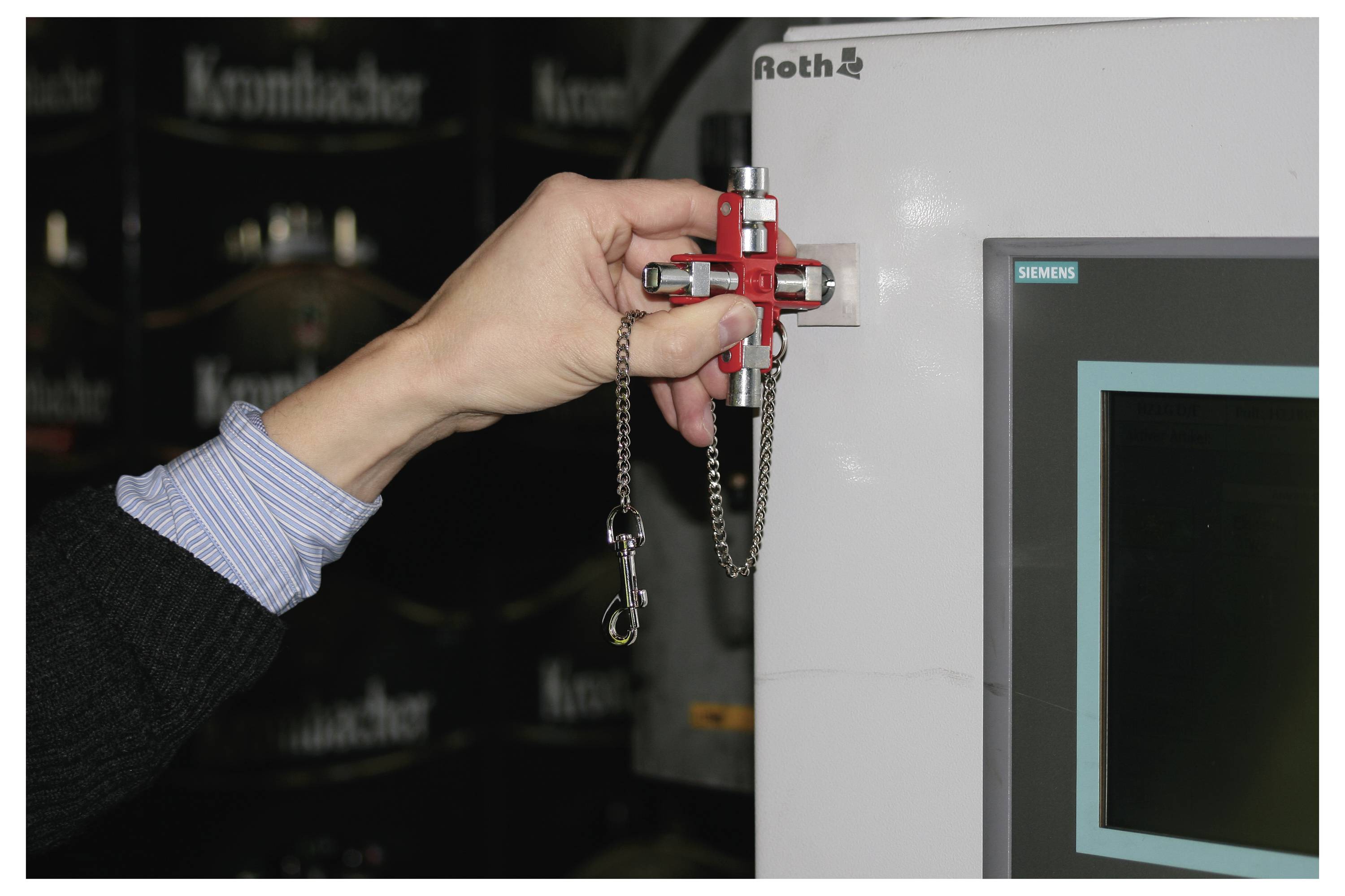 Osoba trzymająca czerwone urządzenie z łańcuchem w pobliżu ekranu monitora w środowisku przemysłowym, wskazująca na techniczną regulację lub konserwację.