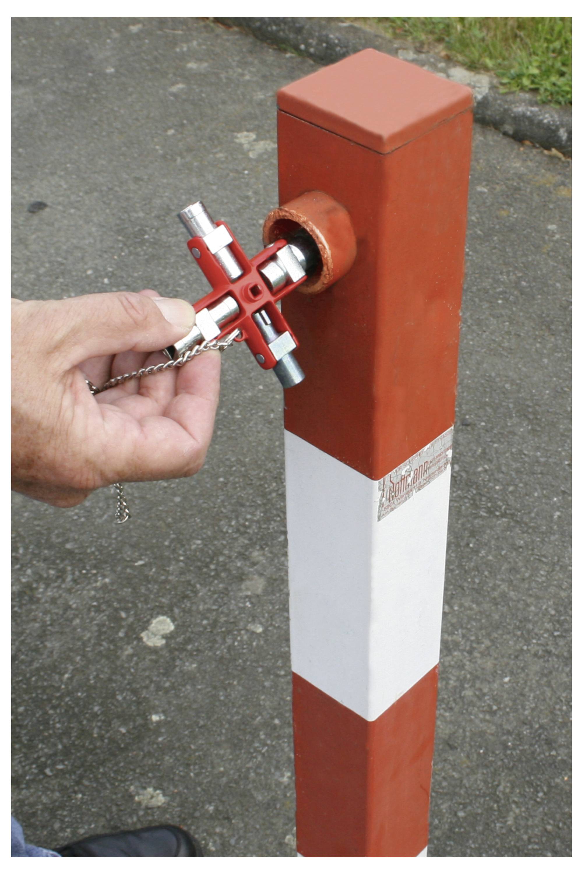 Ręka używa klucza w kształcie krzyża, aby odblokować czerwono-białą zaporę na utwardzonej nawierzchni.