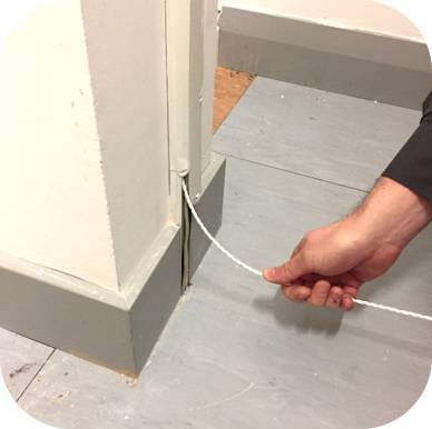 Ręka trzyma białą wtyczkę, która biegnie wzdłuż ściany, obok białej listwy na szarym podłożu.