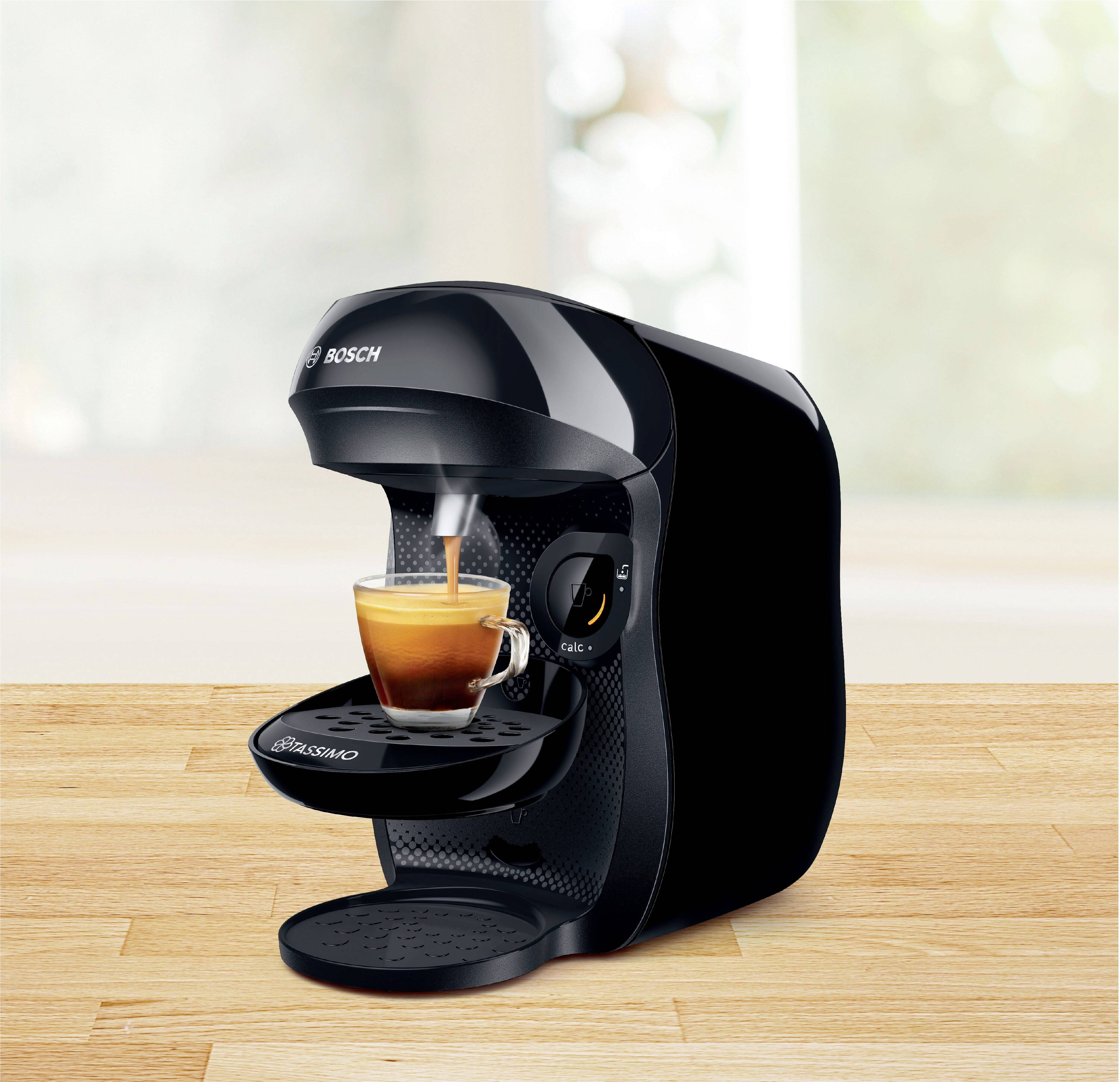 Czarna ekspres do kawy parzy espresso w szklanej filiżance na drewnianym stole. W tle widać rozmyty, jasny pokój.