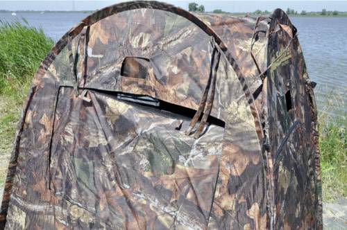 Namiot maskujący nad brzegiem jeziora, otoczony trawą i wodą. Namiot posiada wzór liściasty, który pozwala na skuteczne wtopienie się w krajobraz.