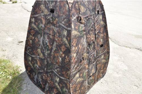 Maskujący parawan myśliwski stoi na betonowej powierzchni. Posiada wzór listowia i gałęzi, służący do maskowania podczas polowania.