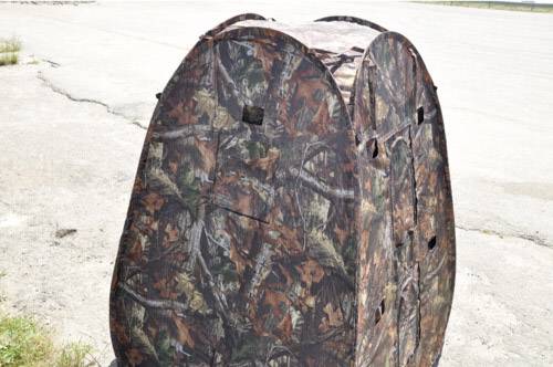 Przenośna kurtyna maskująca z wzorem liści i gałęzi stoi na terenie asfaltowym, przydatna do polowania lub obserwacji zwierząt.