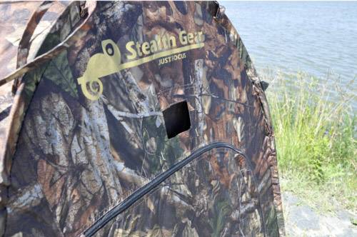 Maskujący namiot Stealth Gear stoi nad brzegiem zbiornika wodnego. W tle widoczne są trawy.