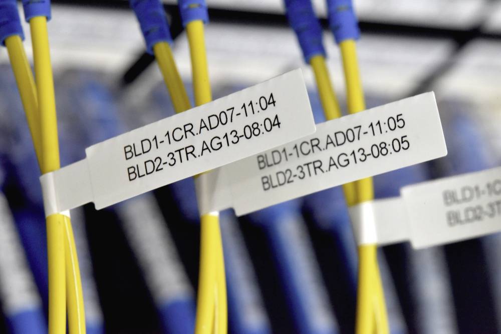 Zbliżenie żółtych kabli światłowodowych z etykietami. Etykiety przedstawiają kody numeryczne wskazujące szczegóły połączeń. Kable są umieszczone w serwerowni.