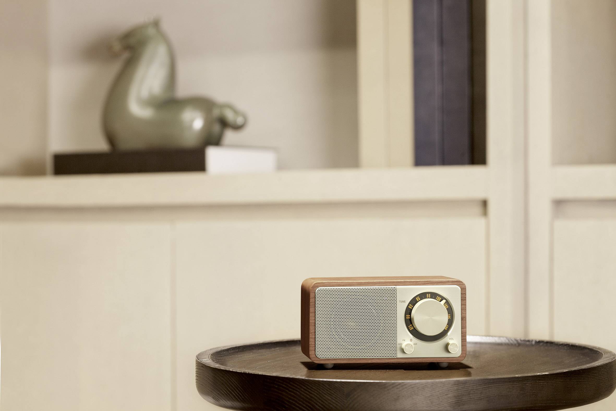 Mały radioodbiornik w stylu retro stojący na drewnianym stole przed białą półką z dekoracyjnym wazonem w tle.