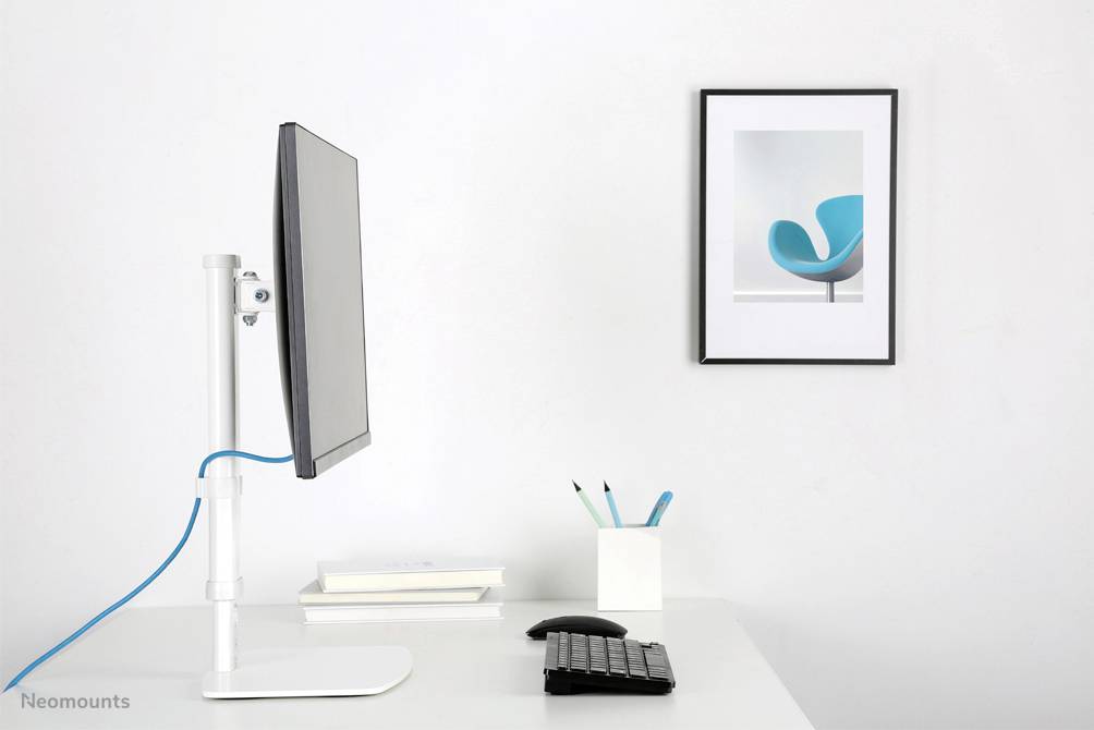 Nowoczesne stanowisko pracy z monitorem zamontowanym z boku białego stołu. Obok leżą książki, długopisy i klawiatura. Na ścianie wisi obraz niebieskiego krzesła.