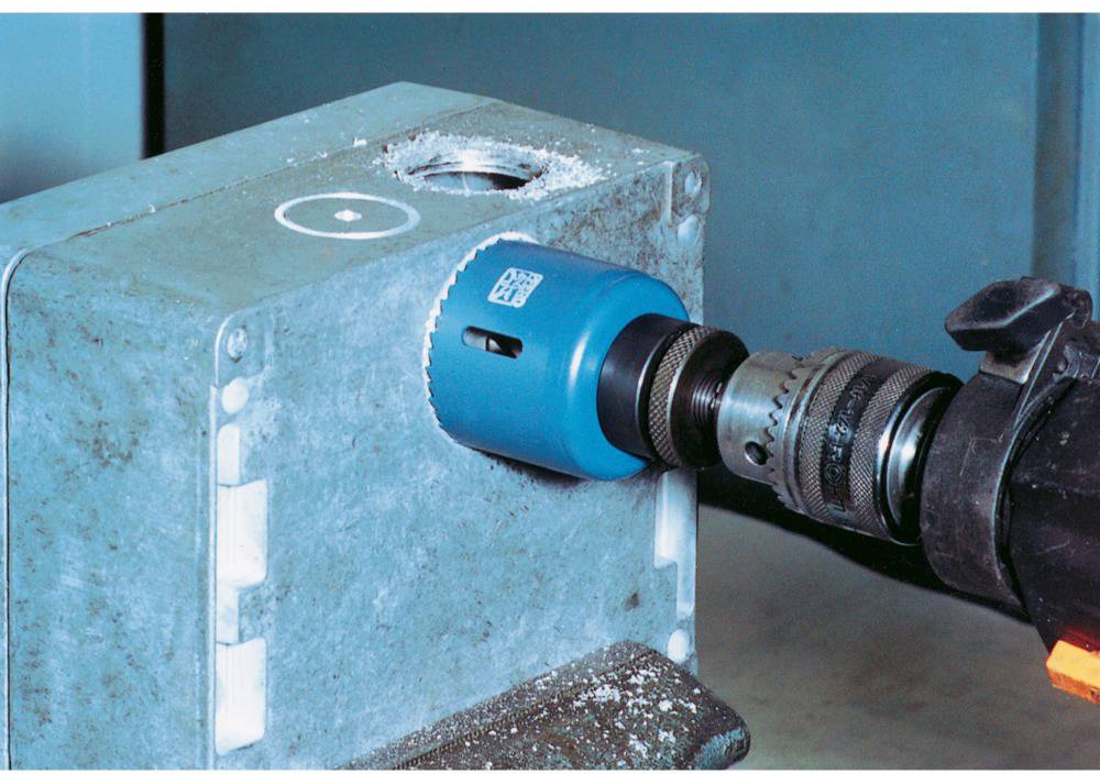 Niebieska piła otworowa wycina okrągły otwór w szarą metalową obudowę. Czarna wiertarka jest przymocowana do piły.