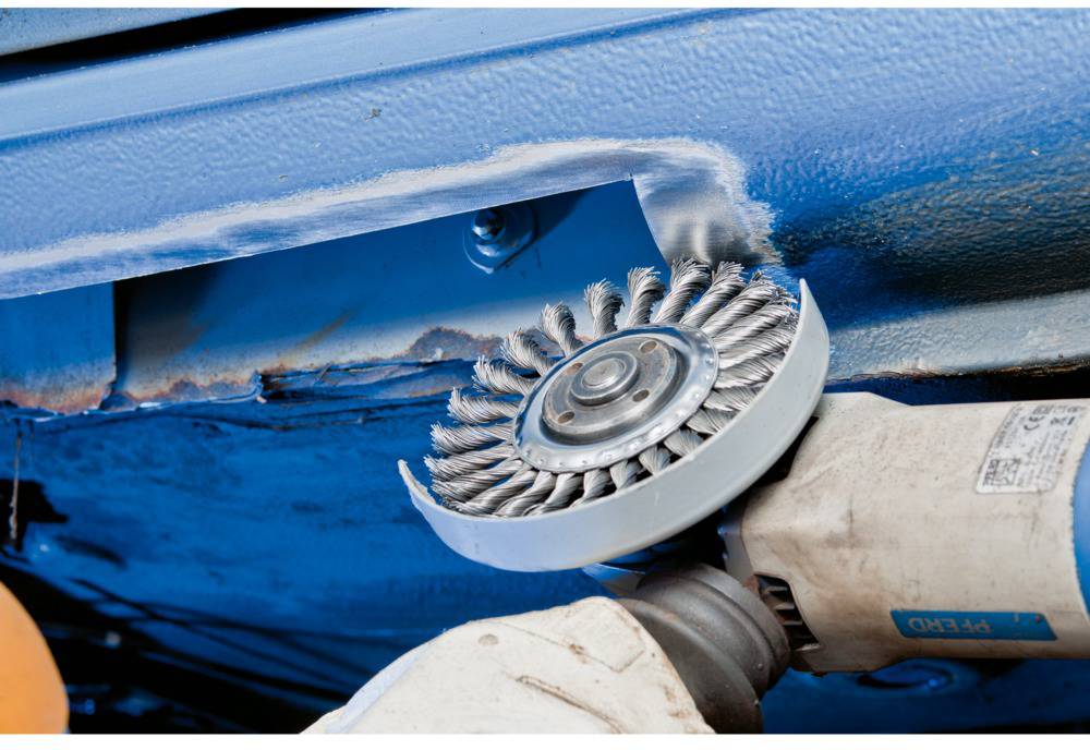 Maszyna z druciakową szczotką szlifuje niebieską powierzchnię metalową. Fokus na procesie czyszczenia, kurz jest usuwany. Odpowiednie do czyszczenia przemysłowego.