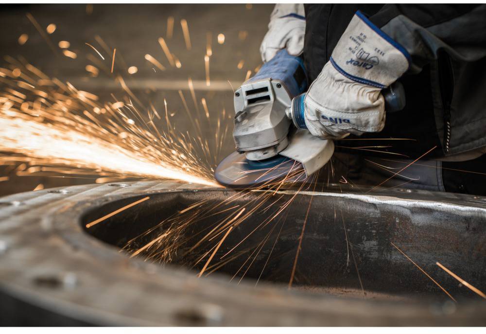 Osoba szlifuje metal szlifierką kątową. Iskry tryskają we wszystkich kierunkach, narzędzie jest trzymane w ochronnych rękawicach.