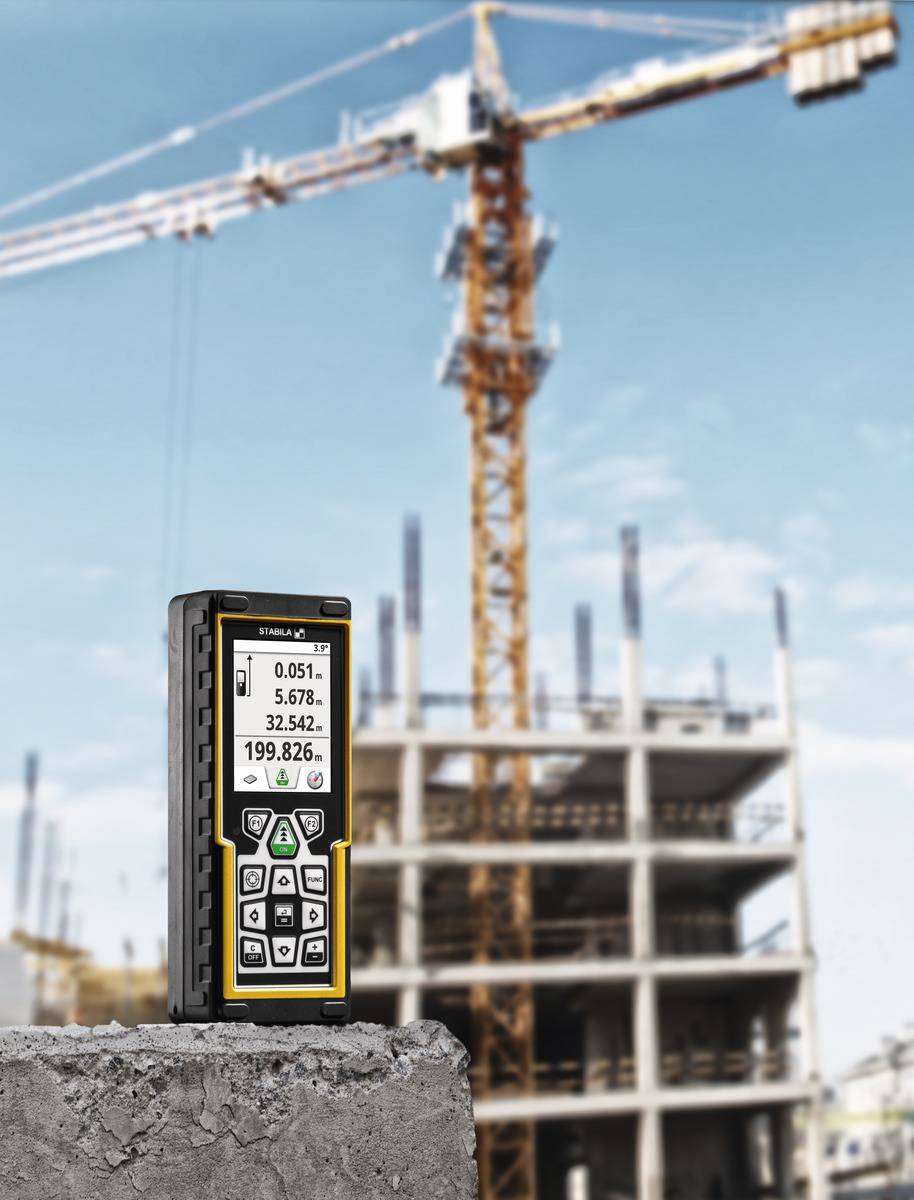 Miernik laserowy leży na betonowej powierzchni, w tle znajduje się budynek w trakcie budowy oraz dźwig budowlany.
