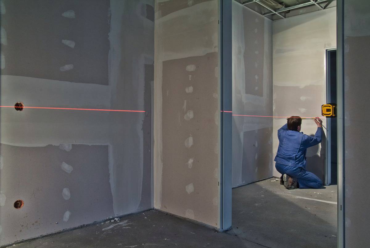 Jedna osoba mierzy w niedokończonym pomieszczeniu z ściankami z płyt gipsowo-kartonowych za pomocą narzędzia laserowego. Czerwone światło lasera przebiega poziomo.