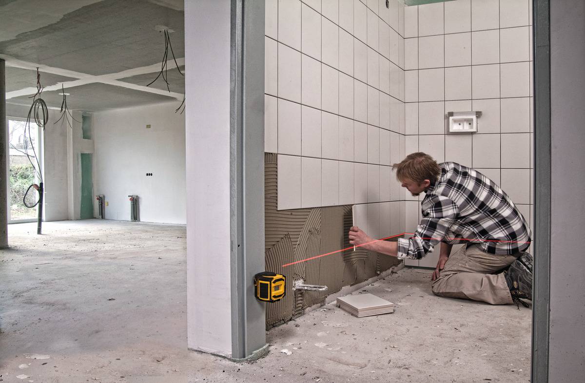 Rzemieślnik klęczy w niedokończonym pomieszczeniu i układa białe kafelki na ścianie. Urządzenie laserowe pomaga w precyzyjnym wyrównaniu.