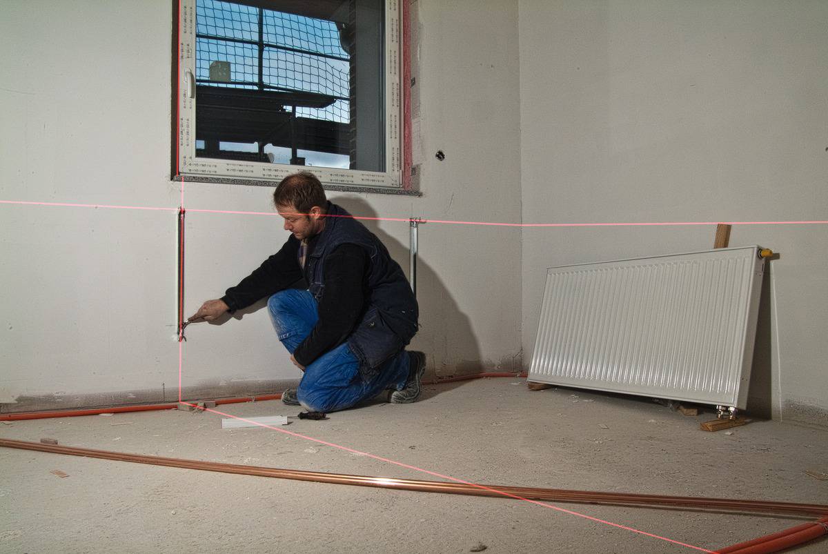 Rzemieślnik klęczy w niedokończonym pomieszczeniu i prowadzi rury miedziane przy pomocy lasera niwelacyjnego. Grzejnik spoczywa oparty o ścianę.