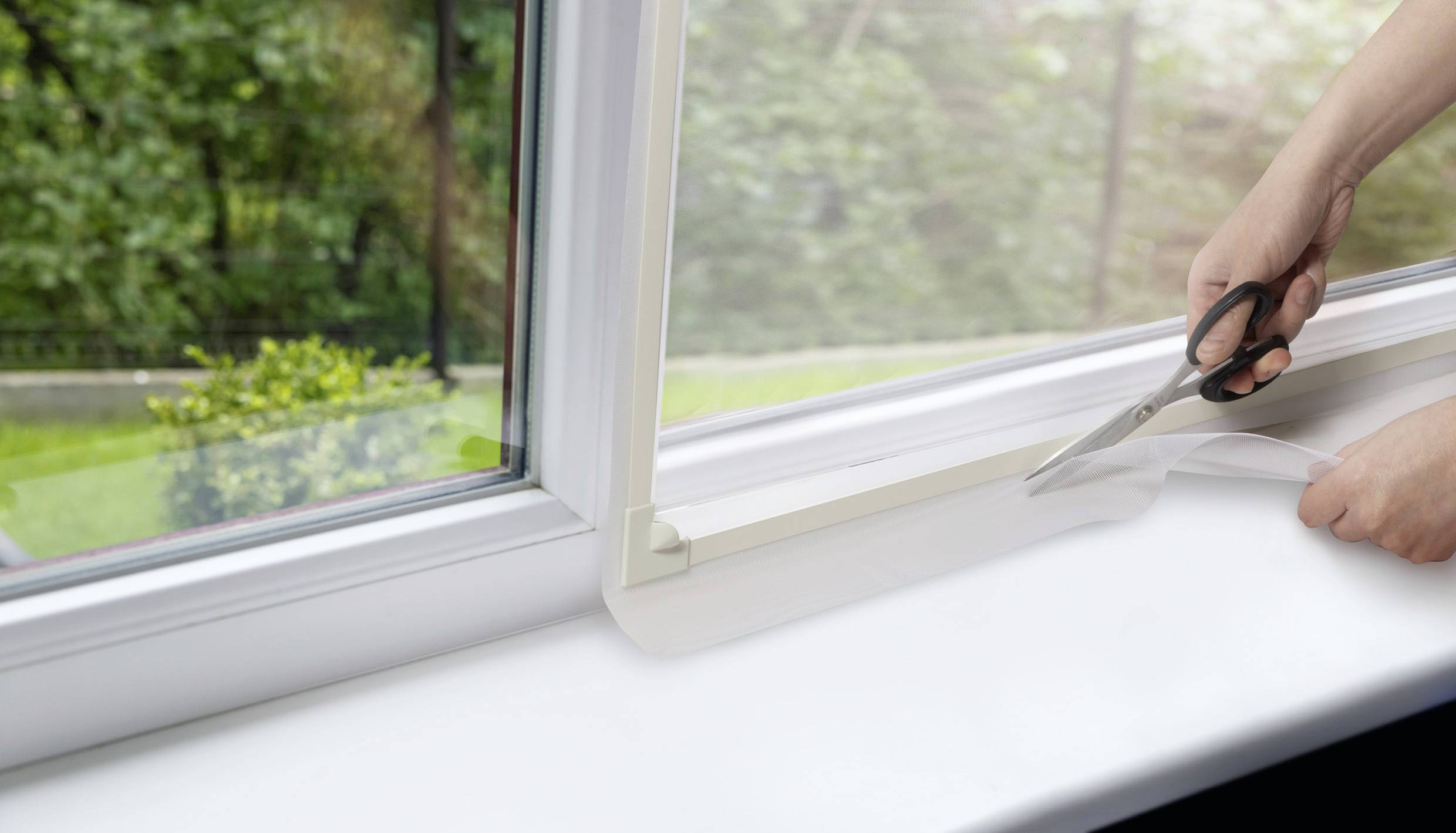 Osoba przycina przeźroczystą folię ochronną wzdłuż ramy okiennej. W tle widać ogród.