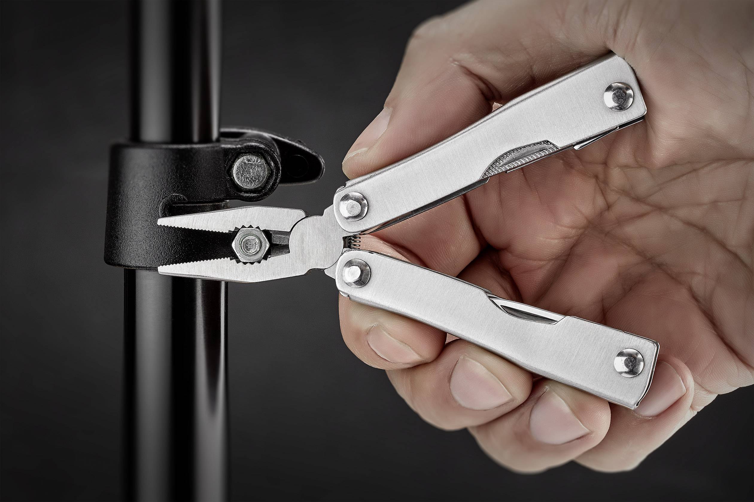 Dłoń trzyma wielofunkcyjne narzędzie z szczypcami, które jest używane przy czarnym pręcie, prawdopodobnie do mocowania lub regulacji.