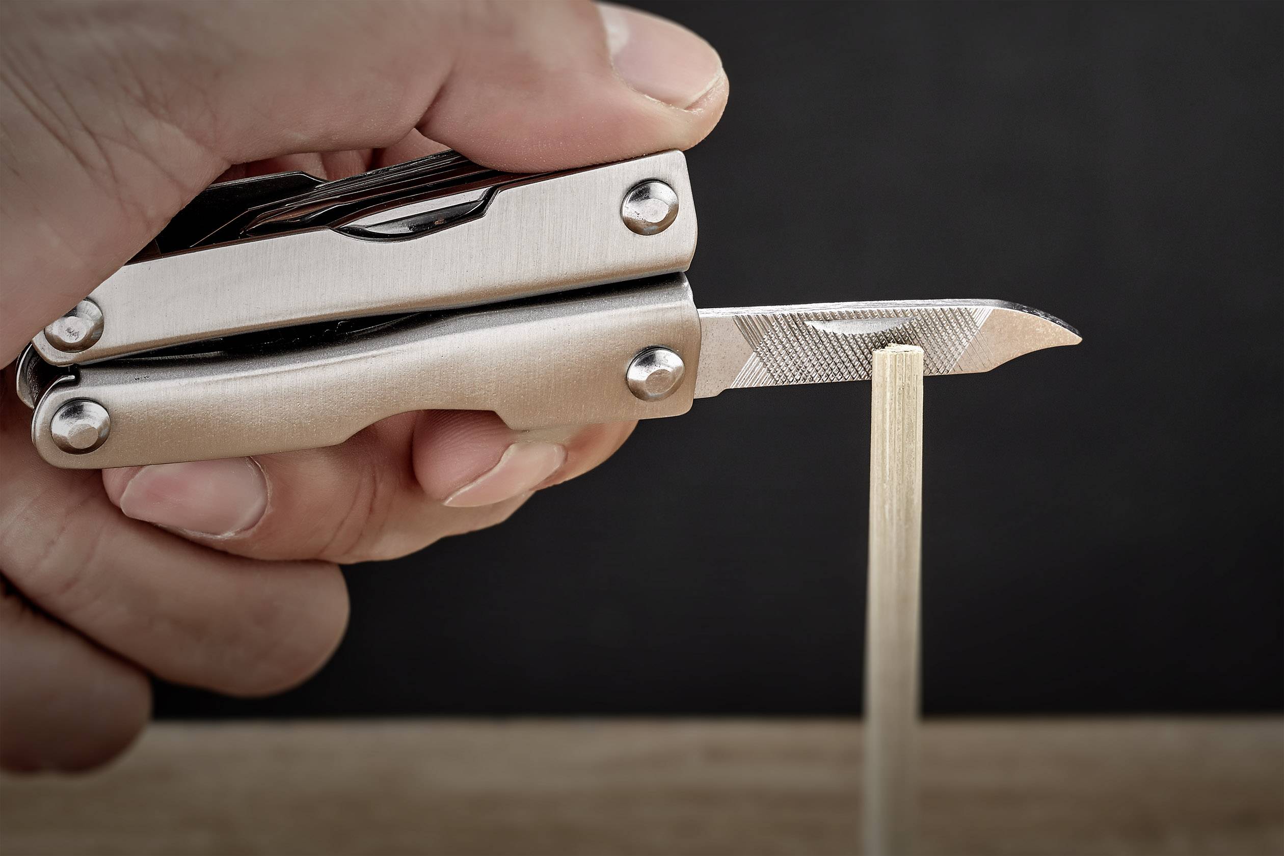 Dłoń trzyma multitool z rozłożonym ostrzem, które zbliża się do pionowo stojącego drewnianego kija.