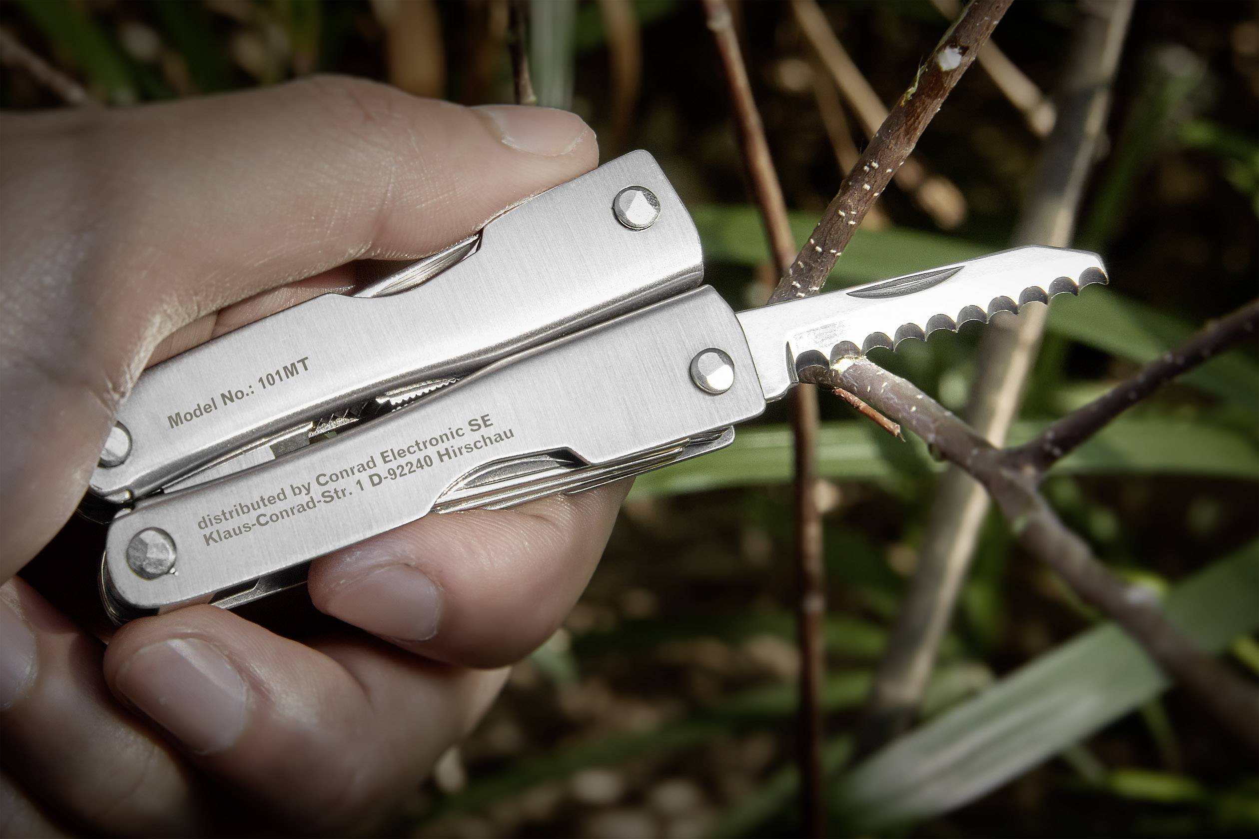 Ręka trzyma wielofunkcyjne narzędzie z rozłożonym ząbkowanym ostrzem, otoczone gałęziami i liśćmi na wolnym powietrzu.