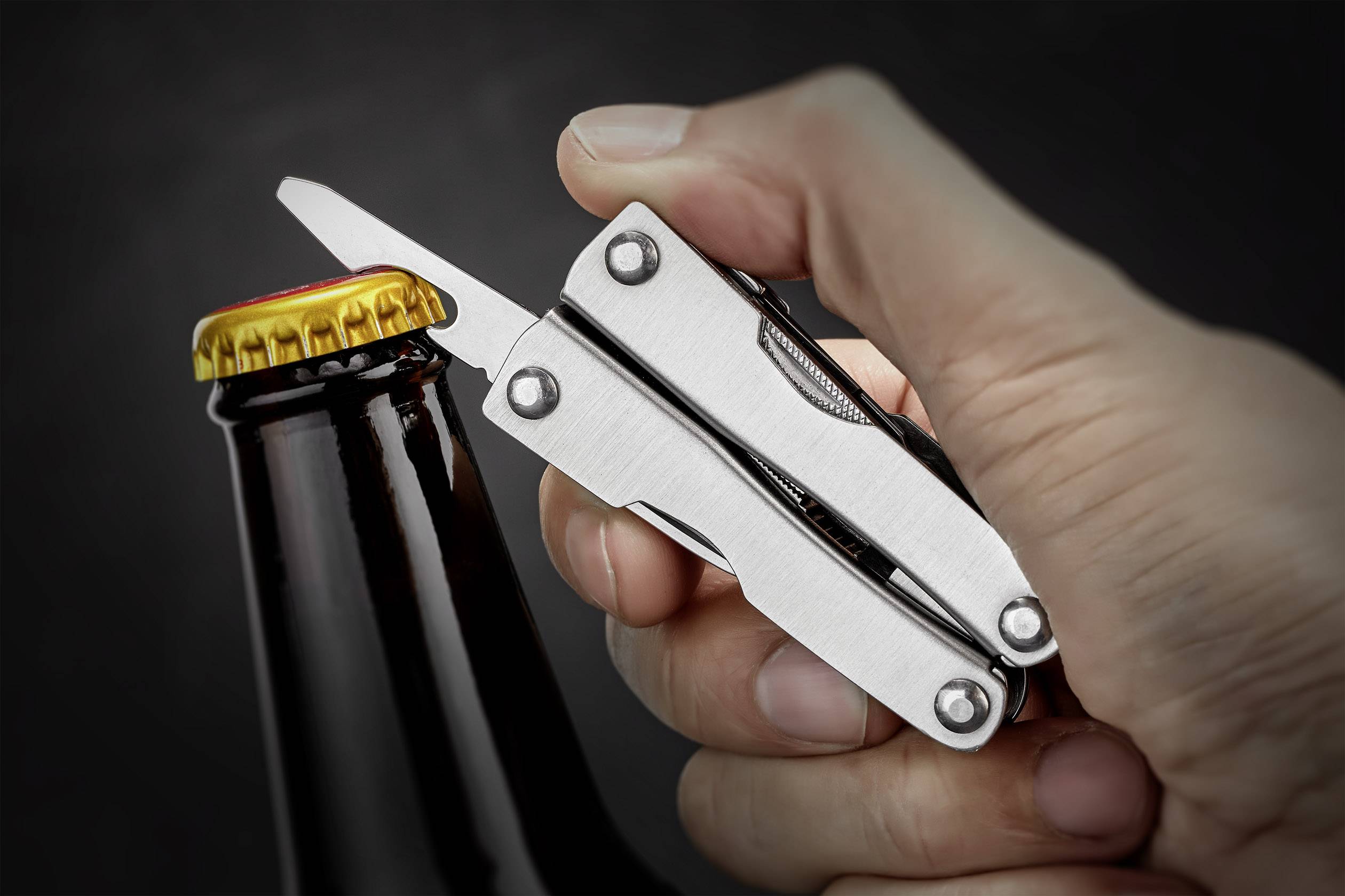 Ręka otwiera butelkę piwa wielofunkcyjnym narzędziem na ciemnym tle.