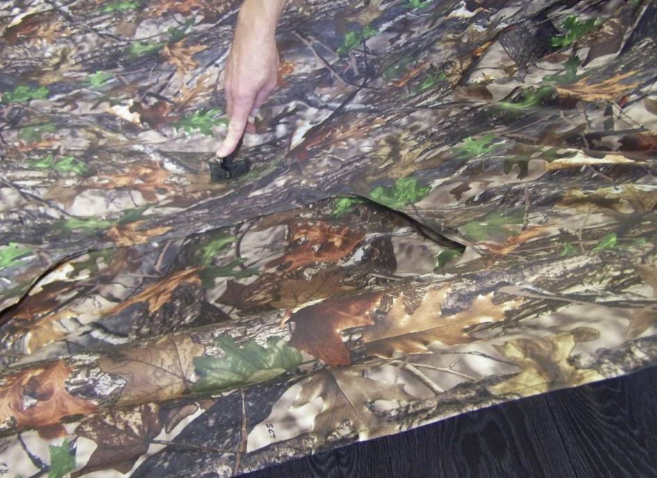 Dłoń wskazuje na szczegół siatki maskującej o wzorze liści i kory drzewa, leżącej na ciemnej drewnianej podłodze.