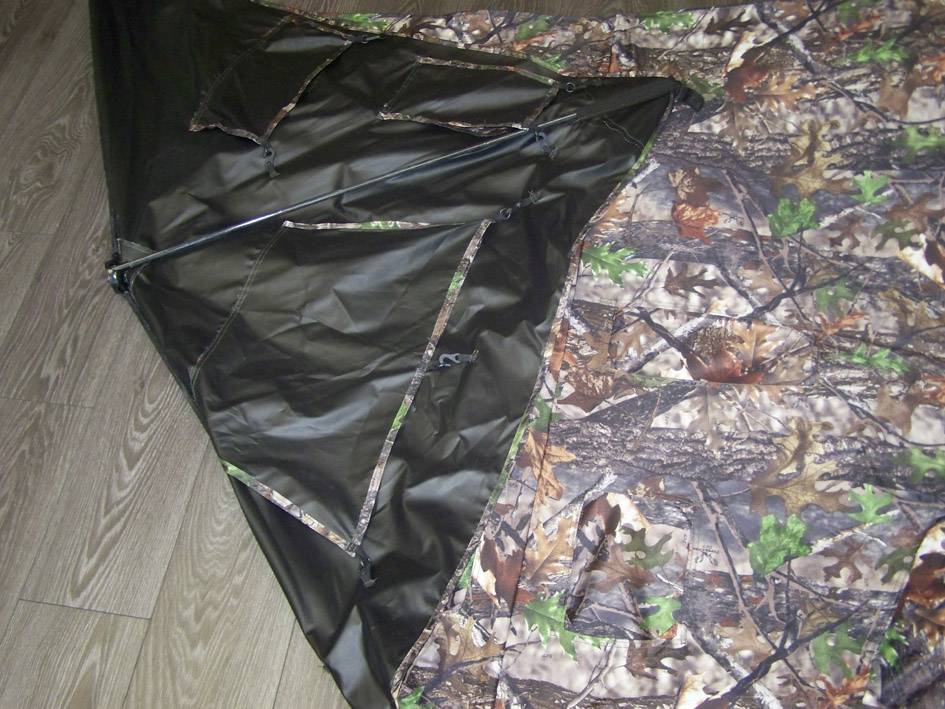 Namiot w kamuflażowym wzorze leży na drewnianej podłodze. Czarny, błyszczący element materiału prezentuje zamkniętą suwaczkę.