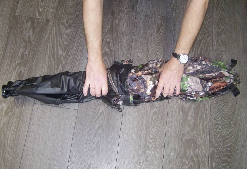 Osoba składa namiot maskujący na szarym drewnianym podłożu. Widoczne są ręce manipulujące materiałem namiotu.