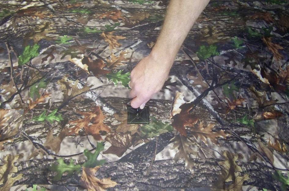 Ręka rozsuwa zamek błyskawiczny na tkaninie w kamuflażowy wzór. Tkanina przedstawia leśny wzór z liśćmi i gałęziami.