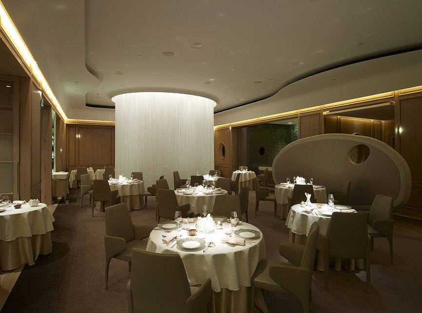 Elegancka restauracja z nakrytymi stołami, dużą centralną lamp oraz przytulnym, stonowanym oświetleniem.