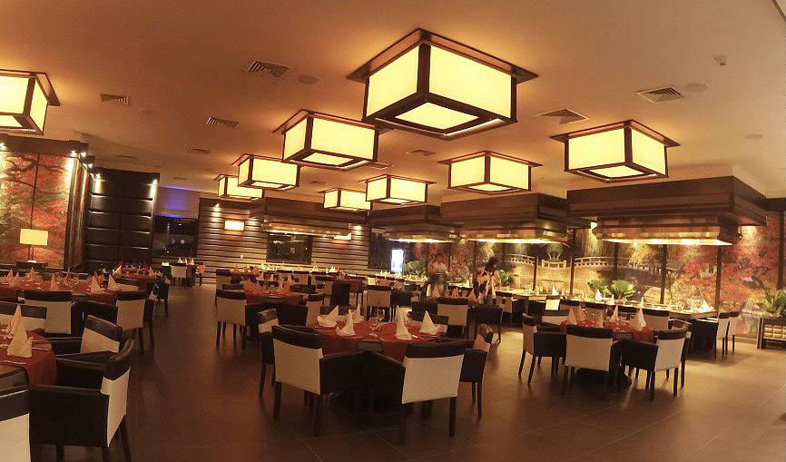 Duża, elegancko oświetlona restauracja z nakrytymi stołami, białymi i czarnymi krzesłami oraz jesiennym malowidłem ściennym w tle.