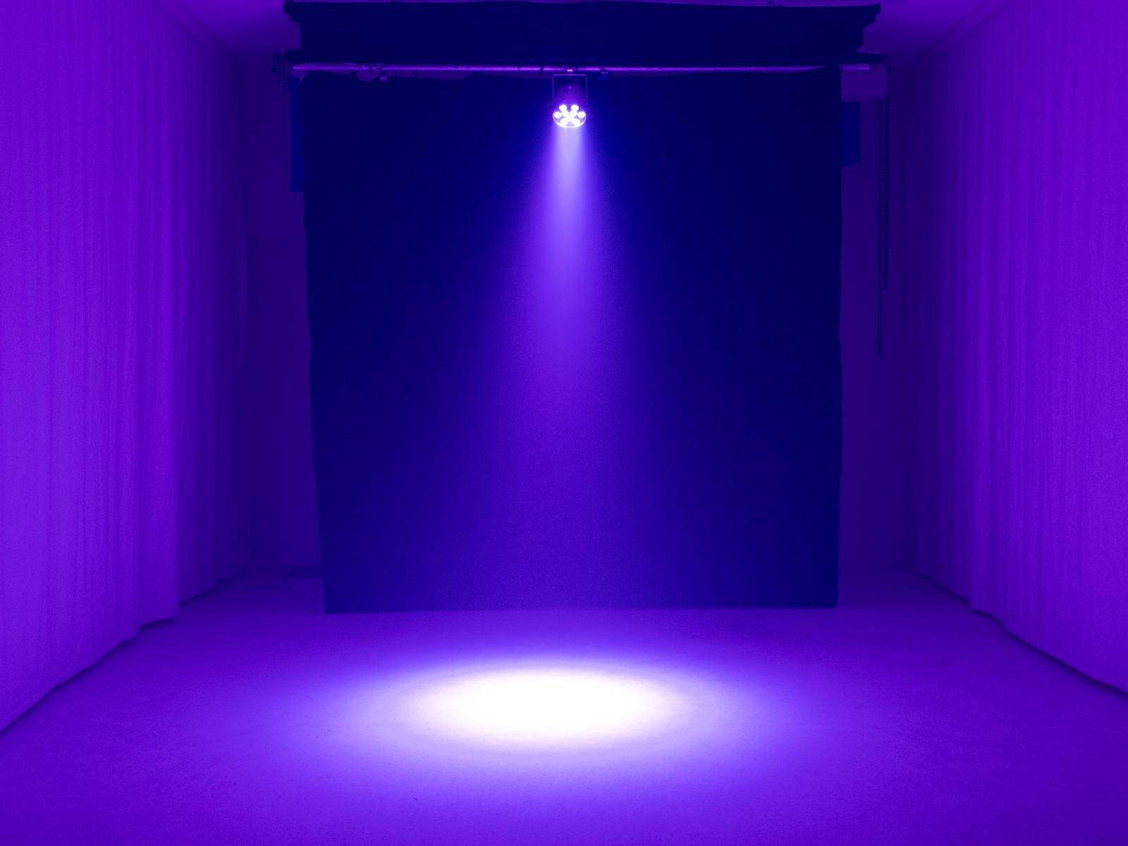 Ciemne pomieszczenie z pojedynczym, skupionym fioletowym reflektorem, który pada na podłogę, tworząc okrągłą płaszczyznę światła.