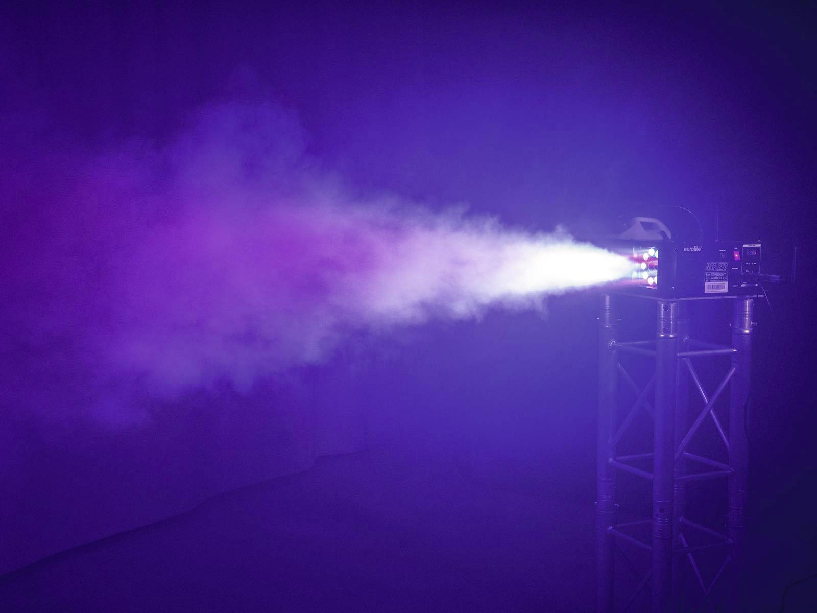 Projektor rzuca wiązkę światła na mgłę, która migocze w różnych kolorach na ciemnym tle.