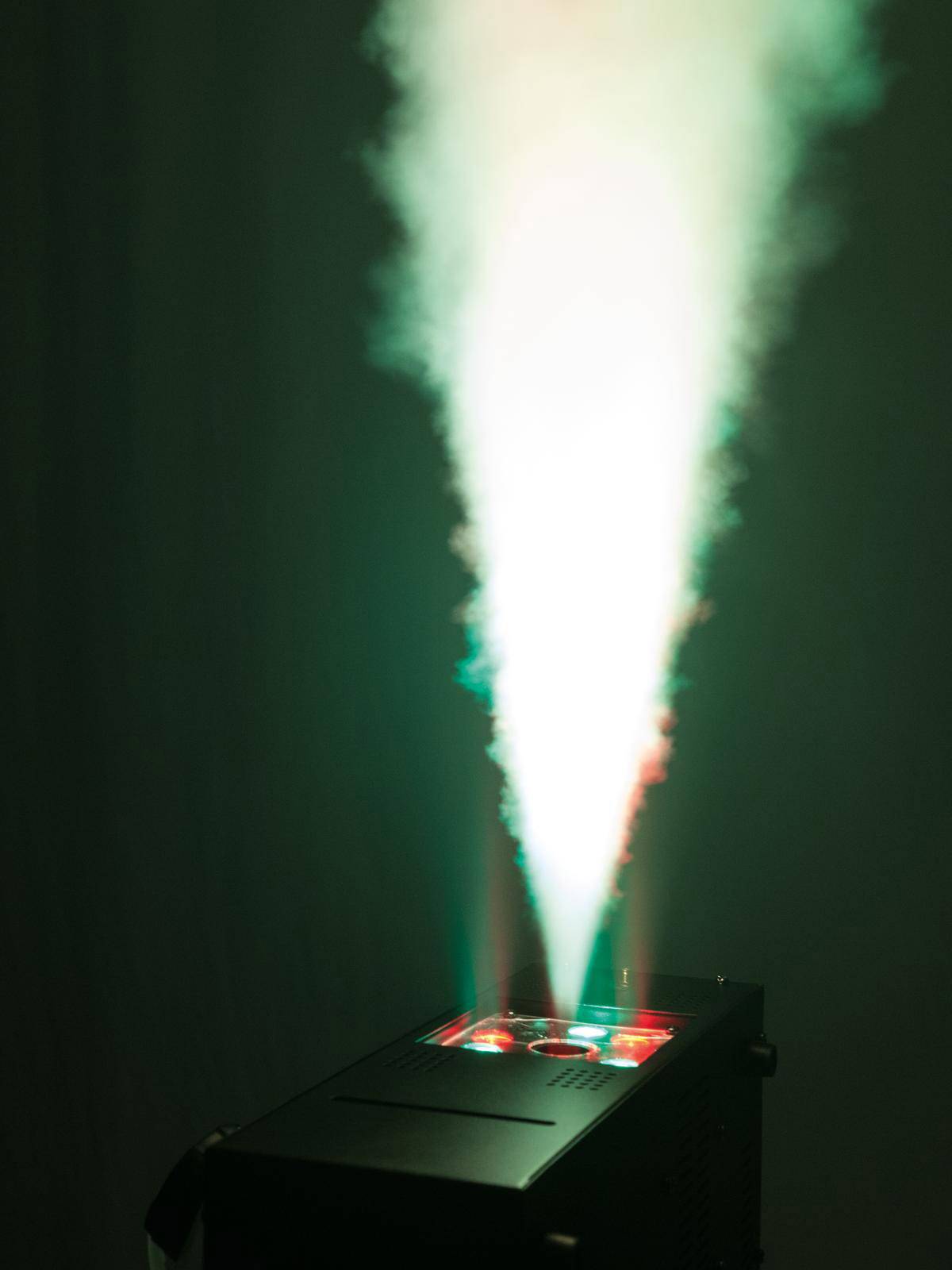 Maszyna mgielna wyrzuca biały snop światła z czerwonymi i zielonymi akcentami w powietrze, na ciemnym tle.
